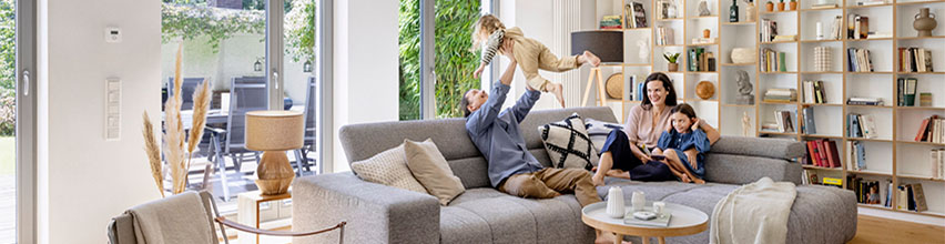 Familj njuter i ljust vardagsrum, pappa lyfter barn, mamma och dotter i grå soffa i modernt smart hem