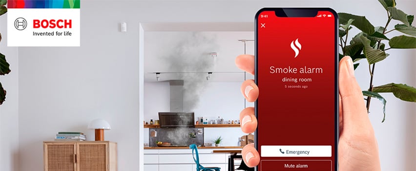 Bosch Smart Home-app visar brandvarnare-avisering från matsal med nödsamtal-knapp och rök från kök i bakgrunden