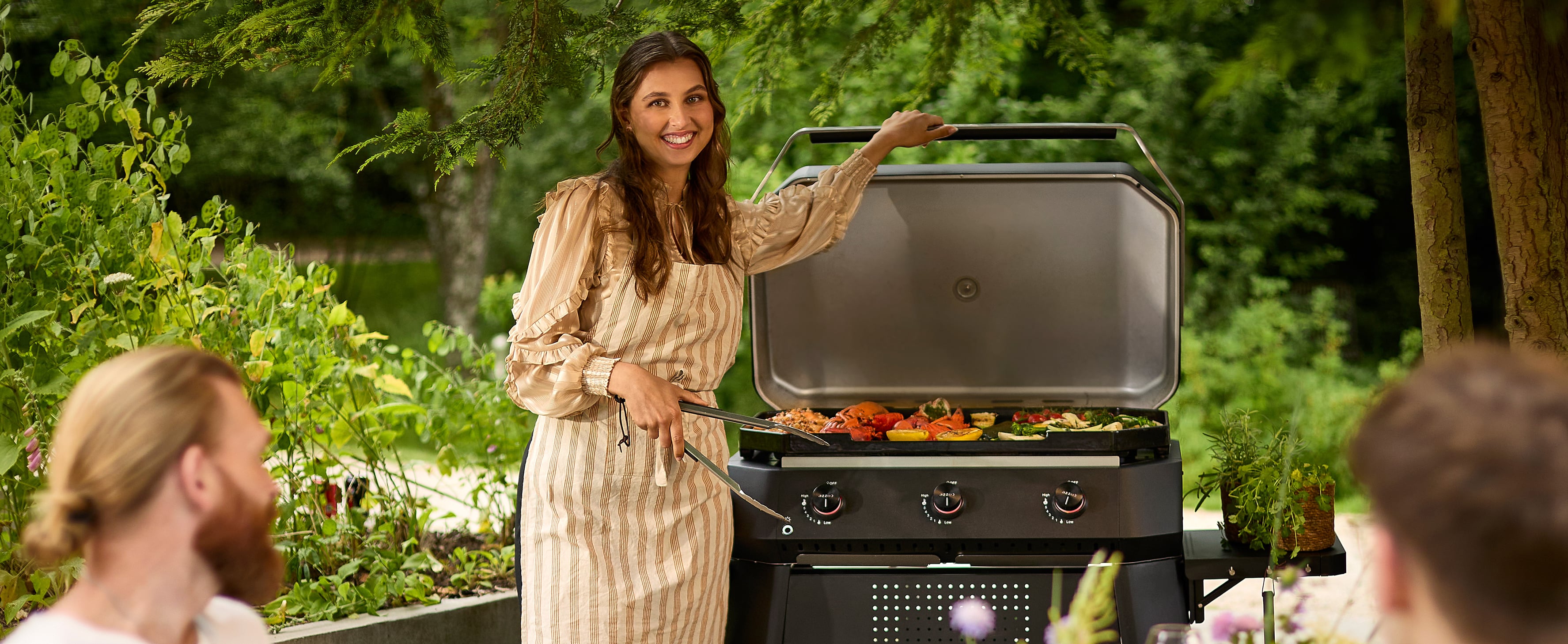 Leende kvinna i förkläde grillar färggranna grönsaker på Cozze gasolgrill med 3 brännare i trädgården – sommarstämning med vänner