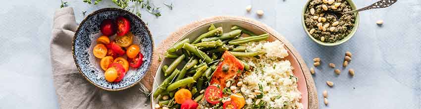 På ett bord står en laxskål med blomkålsris och gröna bönor, toppad med tomater och pesto.