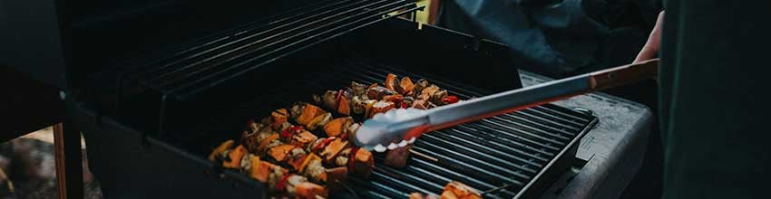 Sju grillade spett med grönsaker vändes med grilltång på en stor, fyrkantig, varm grill.
